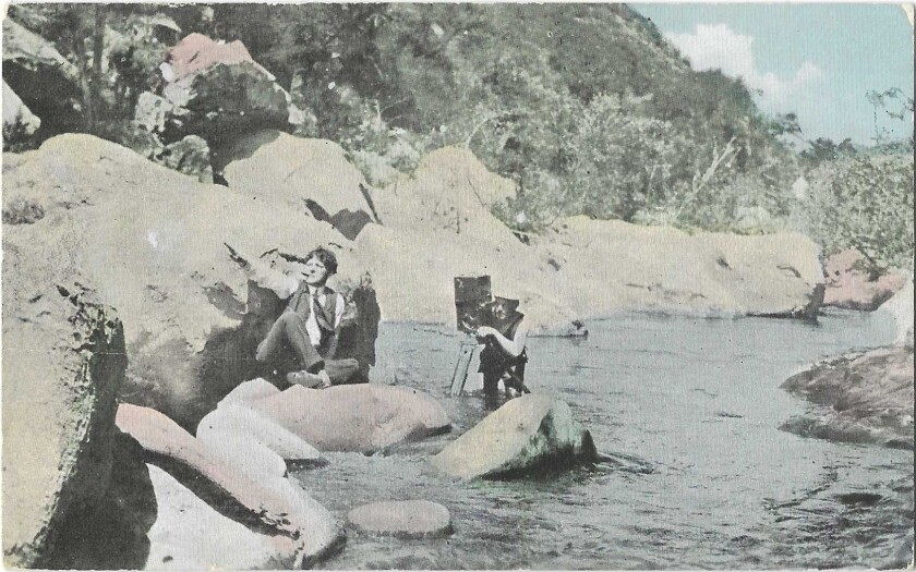 A man with a camera is in the water, while another man sits on a rock and points